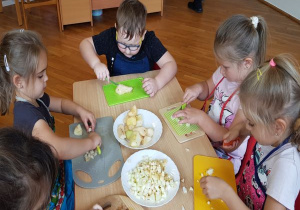 dzieci siedza przy stoliku w fartuszkach i na deskach do krojenia, nożykami plastikowymi kroją obrane owoce na sałatkę